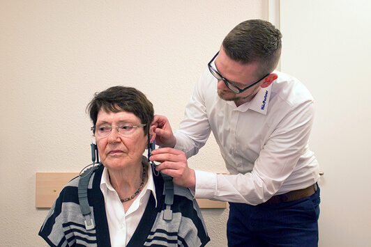 Ein Hörakustiker passt einer älteren Frau ein Hörgerät im Hörtestraum an.